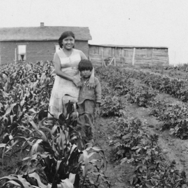 A Ute woman and young boy standing in a crop field with a wooden structure behind them.a