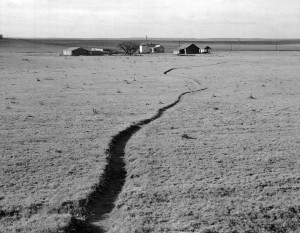The distant structures of a few ranch buildings sit atop a flat landscape. A shallow trench winds from the foreground to the distant buildings.