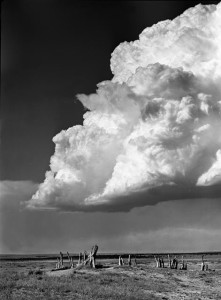 A large storm cloud hangs over a flat landscape. Several pieces of wood protrude from the earth in the foreground.