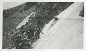 An individual slides through snow down a Clear Creek Basin mountainside. Several men stand at the rocky base of the slope with arms outstretched. 