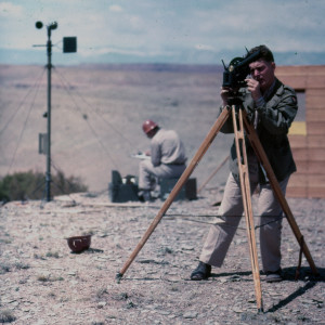 A man adjusts a camera that is positioned on top of a large tripod. He is standing in a desert landscape. Behind him a man wearing a hard hat appears to be writing.