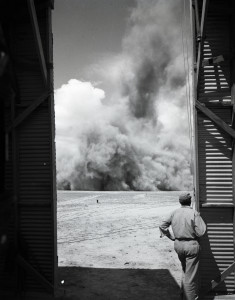 A man wearing a uniform and hat stands at the edge of a large doorway to a barricade and watches an explosion in the not-too-distant background.