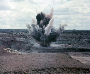 An explosion occurs on top of a small plateau amidst a desert landscape.