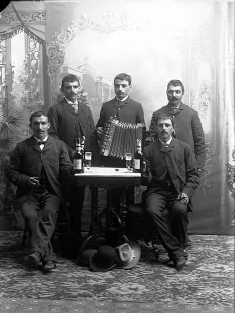 Full-length studio portrait of a group of men, including Peter and Barter, gathered around a table with bottles and glasses of beer. One man holds an accordion.