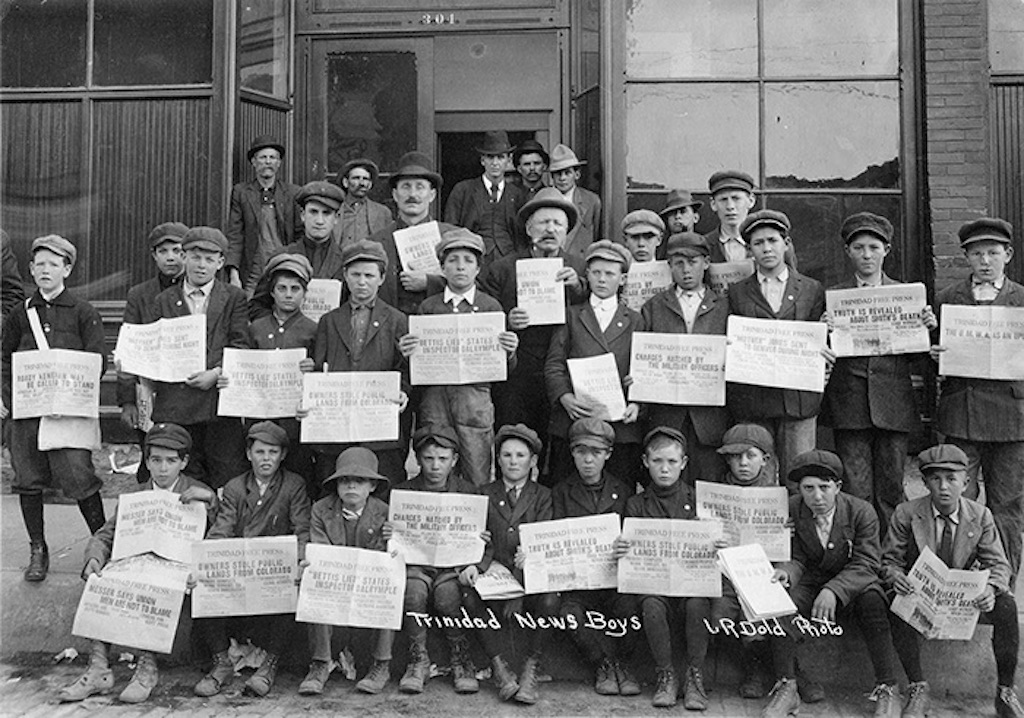 A large group of young boys hold up newspapers showing headlines from the 1914 Ludlow tragedy. 