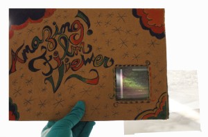 A paperboard sheet displays stylized handwriting which reads "Amazing Film Viewer." In the lower right corner of the paperboard sheet a screened window contains a negative. The screen displays a rainbow, indicating that the negative under investigation is a polyester negative. 