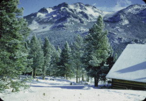 Twin Sisters peaks appear behind a snowy landscape with a wooden structure and pine trees in the foreground.