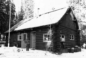 A black and white photo of the cabin with snow on the sloping roof and on the ground below. 
