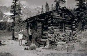 A black and white photo of the cabin with protruding logs on the corners, gabled roof and surrounded by trees. Someone is standing on the left of the cabin and there are prominent mountains in the background on the left. 
