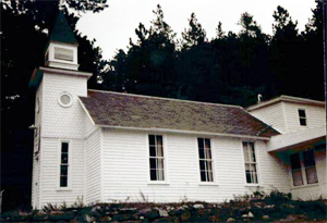 A black and white photo of the side of the white church with steeple on the left and sloping roof on the main section in the center. In the background are dark pine trees.