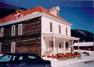 A view of the hotel from an angle, showing a white facade with covered porch and gabled window on hipped roof. On the side are brown boards and white-trimmed windows. 
