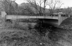 A black and white photo of the bridge with railing on top going over the creek. In the foreground is dead brush and leafless trees and hills stand in the background. 