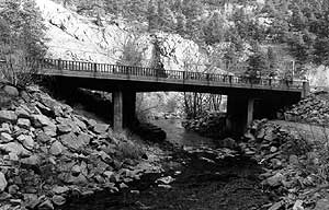 A black and white photo of the bridge with railing crossing over the creek. 