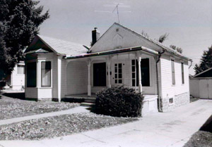 A black and white photo of the house with two gabled roofs and bay window on the left, on the right is a covered porch with the entrance.