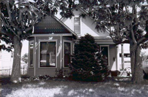 A black and white photo of the house with gabled bay window, and two large trees on either side and shorter evergreen tree in the center.