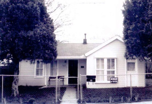A photo of the house from directly in front with chain link fence in front with gabled roof on section on the right and covered porch extending out from the left. On either side stand two leafy trees. 