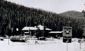 A black and white photo of a snow covered lodge surrounded by a slope of snow covered pine trees and a white field of snow before it.