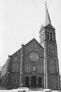 A black and white photo of the church from the front with gabled roof, brickwork, three arched entry, and tall steeple on the right. 
