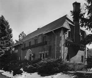 A long angled view of the house in black and white with shrubs and trees in the foreground and sides overhanging snow. On the right of the house is a tall chimney emerging from the second floor. 