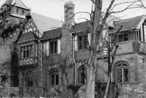 A black and white photo of the elaborately decorated house with lattice work on hall upon the left and chimney in the center. 