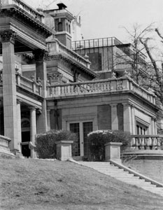 A black and white photo of part of the mansion with terraced balconies, square pillars and stairs leading to entrance. 