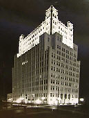 A black and white photo of the building with evenly spaced windows with the upper level well lit and towering on the four corners. 