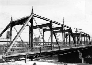A black and white photo of the bridge with double truss and railing on the platform. 