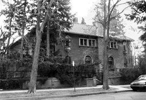 A black and white photo of the house greatly obscured by trees and bushes. 