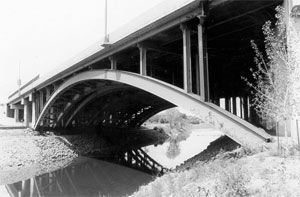 A black and white view from the underside of the bridge, with arches spanning the banks and support beams coming up from them. 