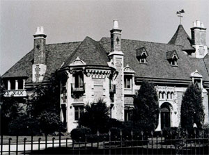 A black and white view of the house beyond a fence, with stone walls and light trim, with gabled windows emerging from the sloped roof. There is a conical roof prominently in the center of the photograph. 