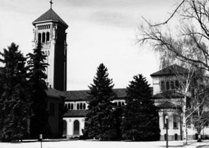 A black and white photo of the campus with tall tower on the left and buildings behind some large evergreens and a large leafless tree on the right. 