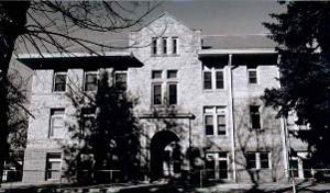 A black and white photo of the building with evergreen trees on the right and center-left. 