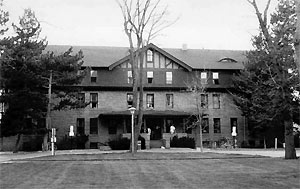 A blacka nd white photo of the hall from across a lawn, with leafless tree in the center and evergreens on either side. 