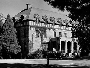 A black and white photo of the building with tall hipped roof and dormer windows protruding. On either side are large trees. 