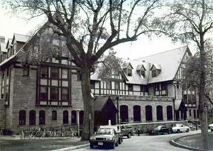 A black and white photo of the hall with with brick base with arcade and tall gabled roof with gabled windows inset and below is lattice work. In the center and right of the picture stand two large leafless trees.