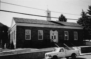A black and white photo of the post office with tall windows framed in white against the brick background. In the center of the top is a small white tower. 