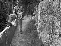 A black and white photo of part of the trail with rock on the right and a person holding a hat and propping their foot on the ledge on the left. 