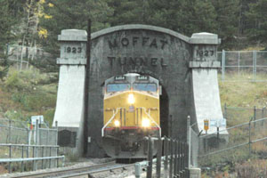 A view of the entrance of the tunnel with a locomotive emerging from it to the tracks bending to the right. 