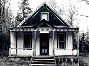 A black and white photo from the front of the building with dark gabled roof and white entrance with covered porch and steps leading to the entrance.