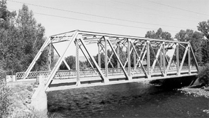 A black and white photo of the bridge running over the river with truss. 