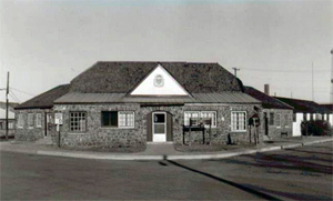 A black and white photo of a building with hipped roof and gable in center over one floor.