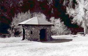 A photo of a small shelter with stone walls and octagonal hipped roof, surrounded by trees in black and white.