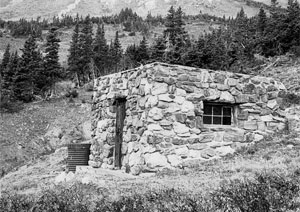 A black and white photo of the building made of stone with rows of evergreen trees and mountains in the background. 