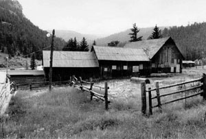 A black and white photo of the ranch surrounded by mountains, with a fence in front.