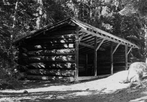 A black and white photo of the stable  with large log walls on the side and sloping overhang before a row of pine trees with snow on the ground before the stables. 