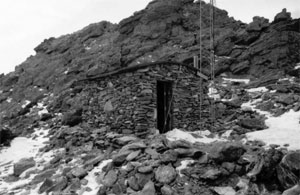 A black and white photo of the shack with tower next to it, and rocky peak in the background, in the foreground there is snow lain about sporadically. 