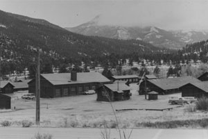 A black and white photo of some buildings in the district with snow lain in the foreground and rolling mountains in the background covered by trees. 