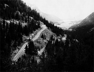 A black and white photo of the road on the side of a mountain in a heavily forested valley with open fields in the distance.