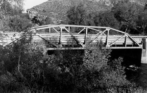 A black and white photo of the bridge with girders on the sides and many trees around. 