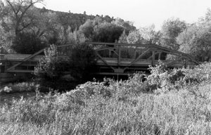 A photo of the bridge from a bank in black and white. The bridge has support girders on either side and lots of foliage around. 